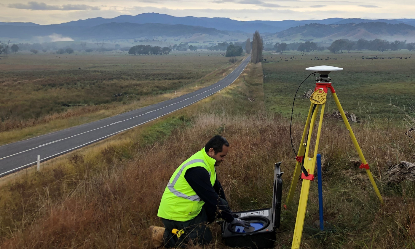 A man in a high visibility jumper sets up surveying equipment next to a rural highway. Mt Kosciuszko is in the background and fenced off fields are on either side of the road.