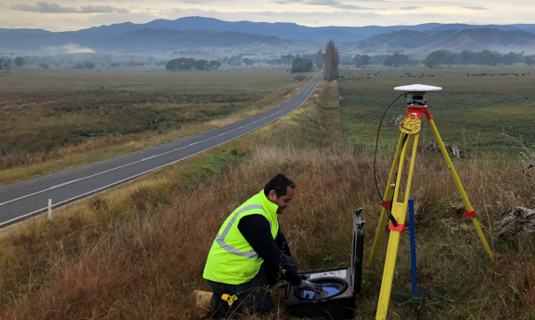 A man in a high visibility jumper sets up surveying equipment next to a rural highway. Mt Kosciuszko is in the background and fenced off fields are on either side of the road.