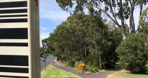 A surveyor and survey equipment standing in front of trees and a suburban street.  
