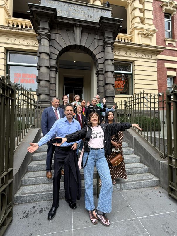 A group of people posing on steps in front of the Queen Victoria Women's Centre, with the building's historic entrance and signage visible behind them. The structure features ornate architecture and iron gates.