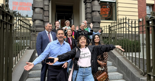 A group of people posing on steps in front of the Queen Victoria Women's Centre, with the building's historic entrance and signage visible behind them. The structure features ornate architecture and iron gates.  