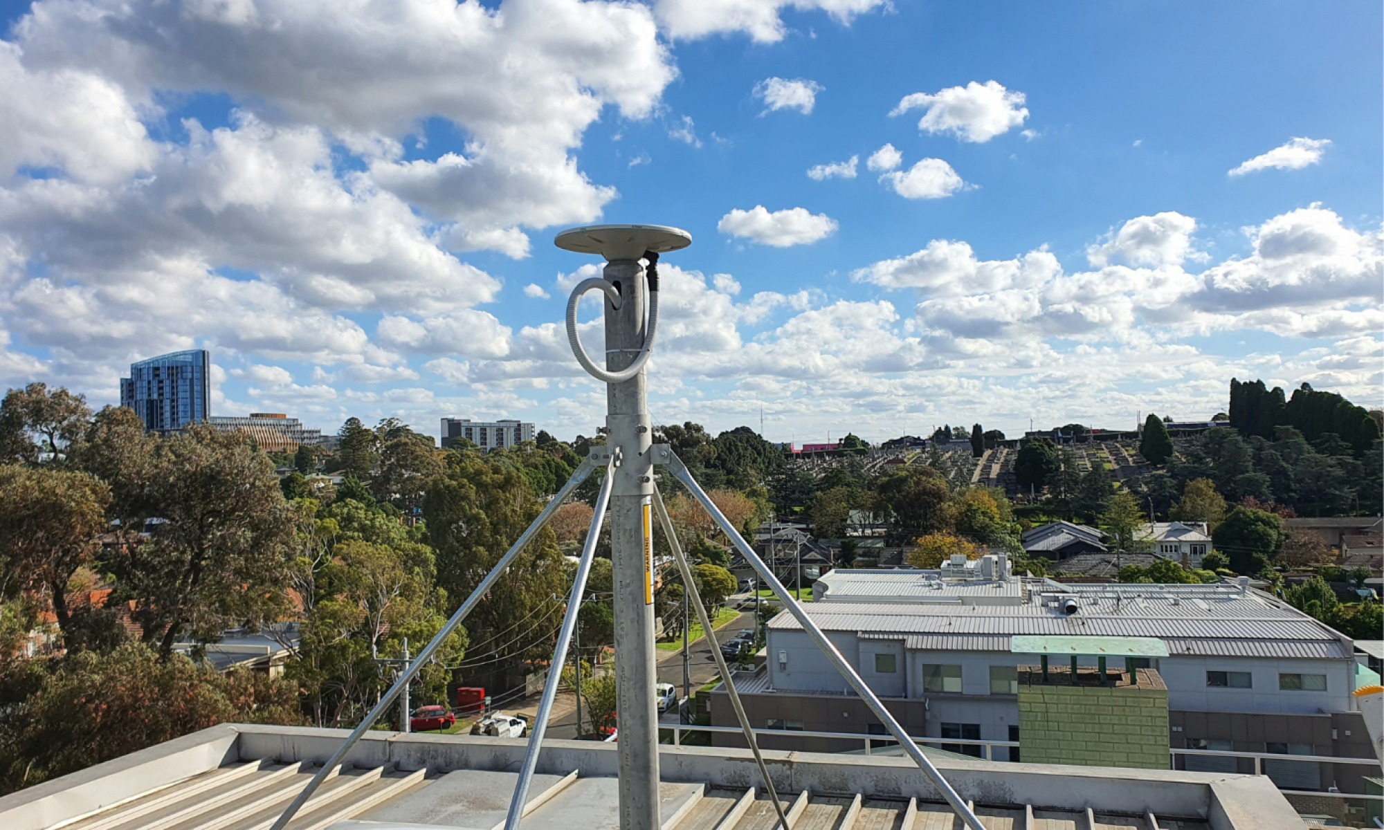 A GPSnet CORS mast and antenna mounted on the roof of a building with trees and houses in the background and a cloudy blue sky.