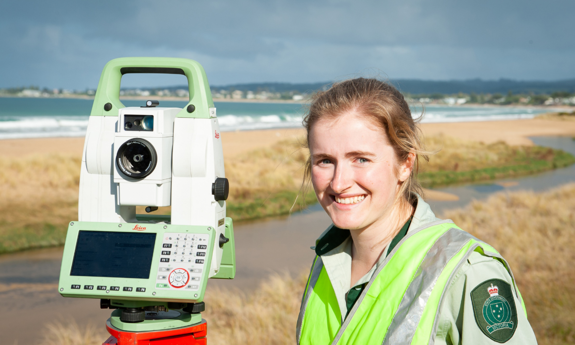 A woman wearing a yellow high-visibility vest stands next to surveying equipment in front of a footpath and breaking waves on a beach.