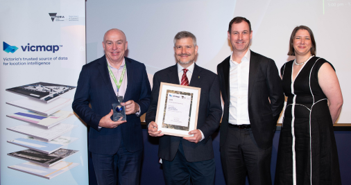Three men and a woman stand in front of a Vicmap banner. Two of the men are holding an award.  
