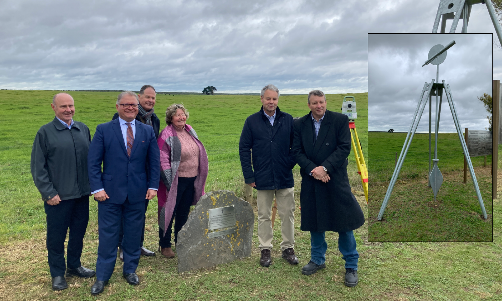 A photo of 5 men and 1 woman standing around and behind a large rock with a metal plaque on it. There is a green field behind them and grey clouds and everyone is smiling. A yellow, red and grey theodolite is positioned behind them with its tripod legs and technical aparatus on top. An inset photograph shows a 3 metre tall steel replica of a theodolite.