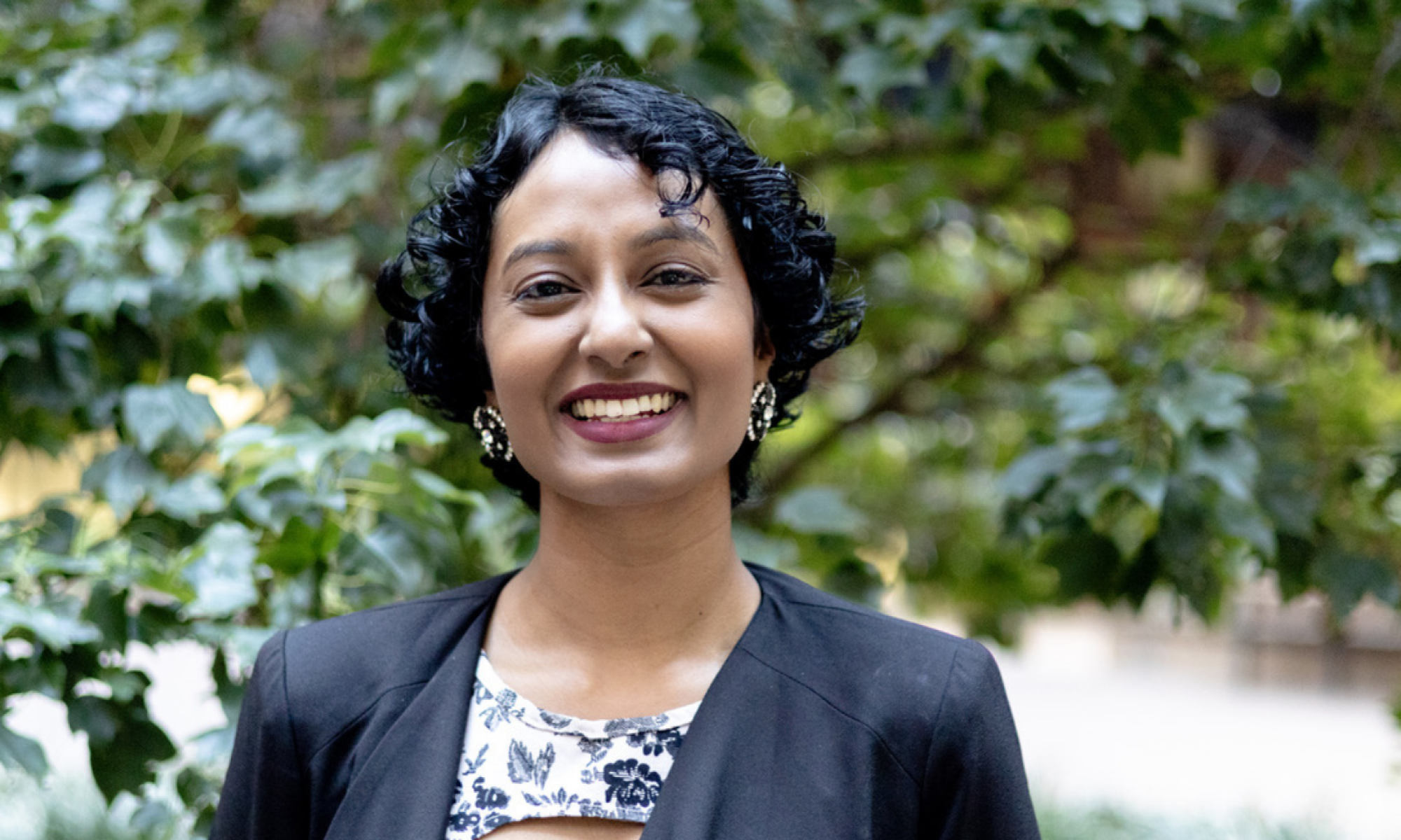 Head-and-shoulders portrait of Shaz Sivanesan standing outdoors with leafy green trees in the background. Shaz is wearing a dark jacket over a patterned top and is facing the camera.