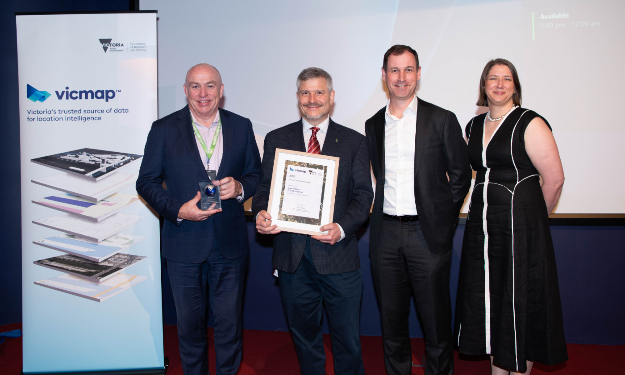 Three men and one woman pose in front of a Vicmap banner. Two of the men are holding an award.