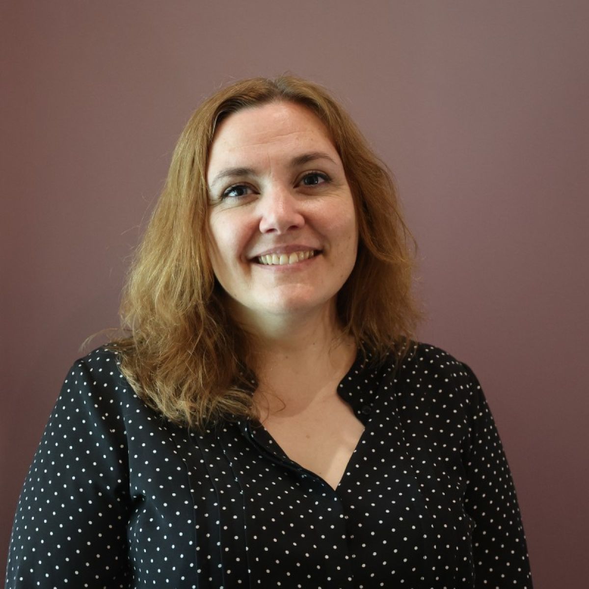 Head-and-shoulders portrait of Melanie Carews standing in front of a mauve-coloured wall. Melanie is wearing a dark blouse with a small white polka-dot pattern and is facing the camera.