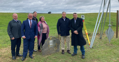 A photo of 5 men and 1 woman standing around and behind a large rock with a metal plaque on it. There is a green field behind them and grey clouds and everyone is smiling. A yellow, red and grey theodolite is positioned behind them with its tripod legs and technical aparatus on top.  