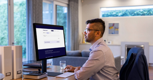 A professionally dressed man sits at a desk looking at a computer monitor with the words 'property search' visible at the top.  