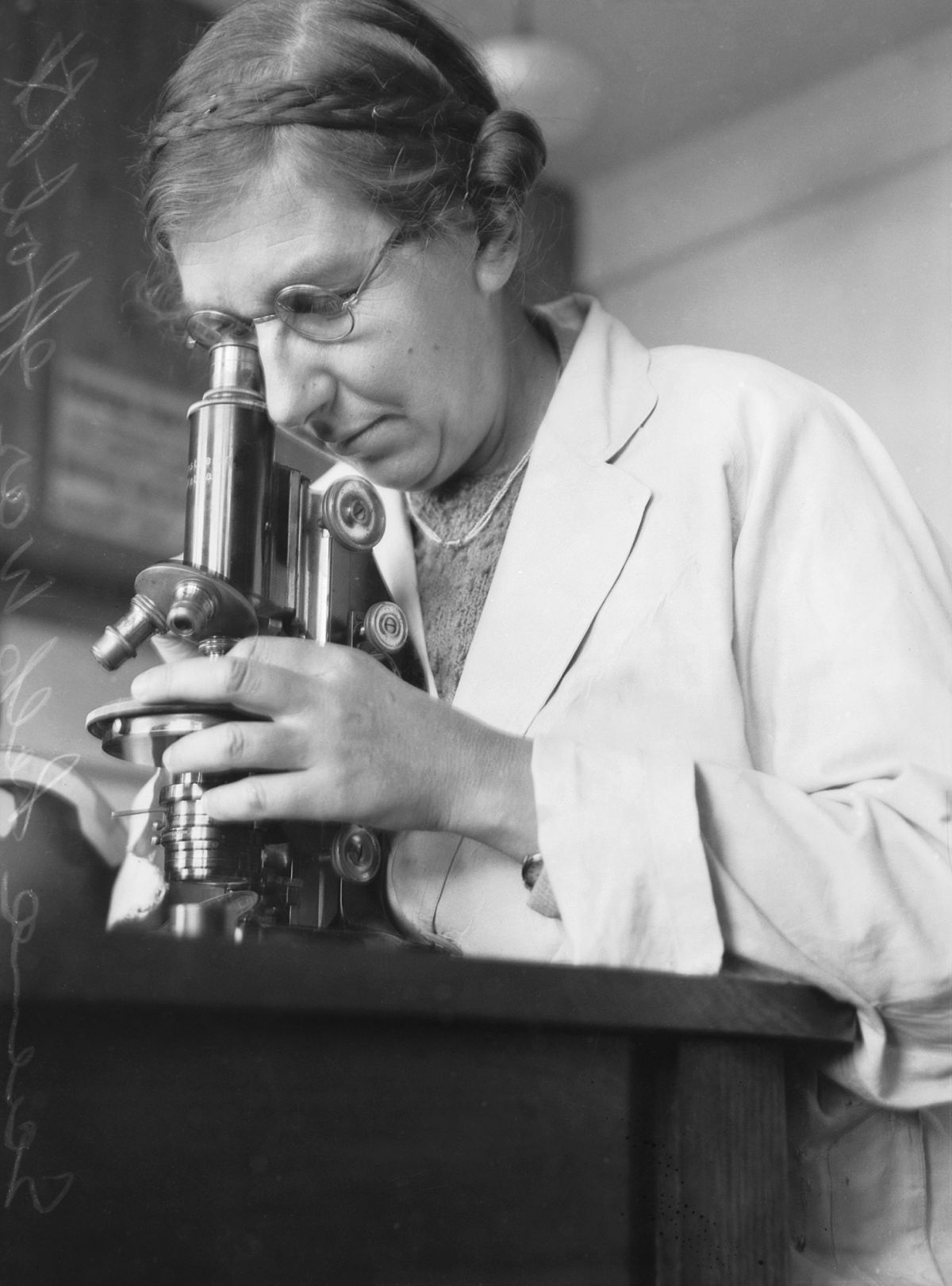 Black-and-white photograph of a Dr Isabel Clifton Cookson in a laboratory coat looking through a microscope while seated at a laboratory bench. The microscope is held with both hands and laboratory equipment is visible in the background.