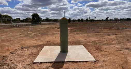 A grey tower installed on a concrete square, surrounded by red dirt. There are trees in the background, the sun is shining and casting shadows, and the sky is blue and dappled with grey and white clouds.  