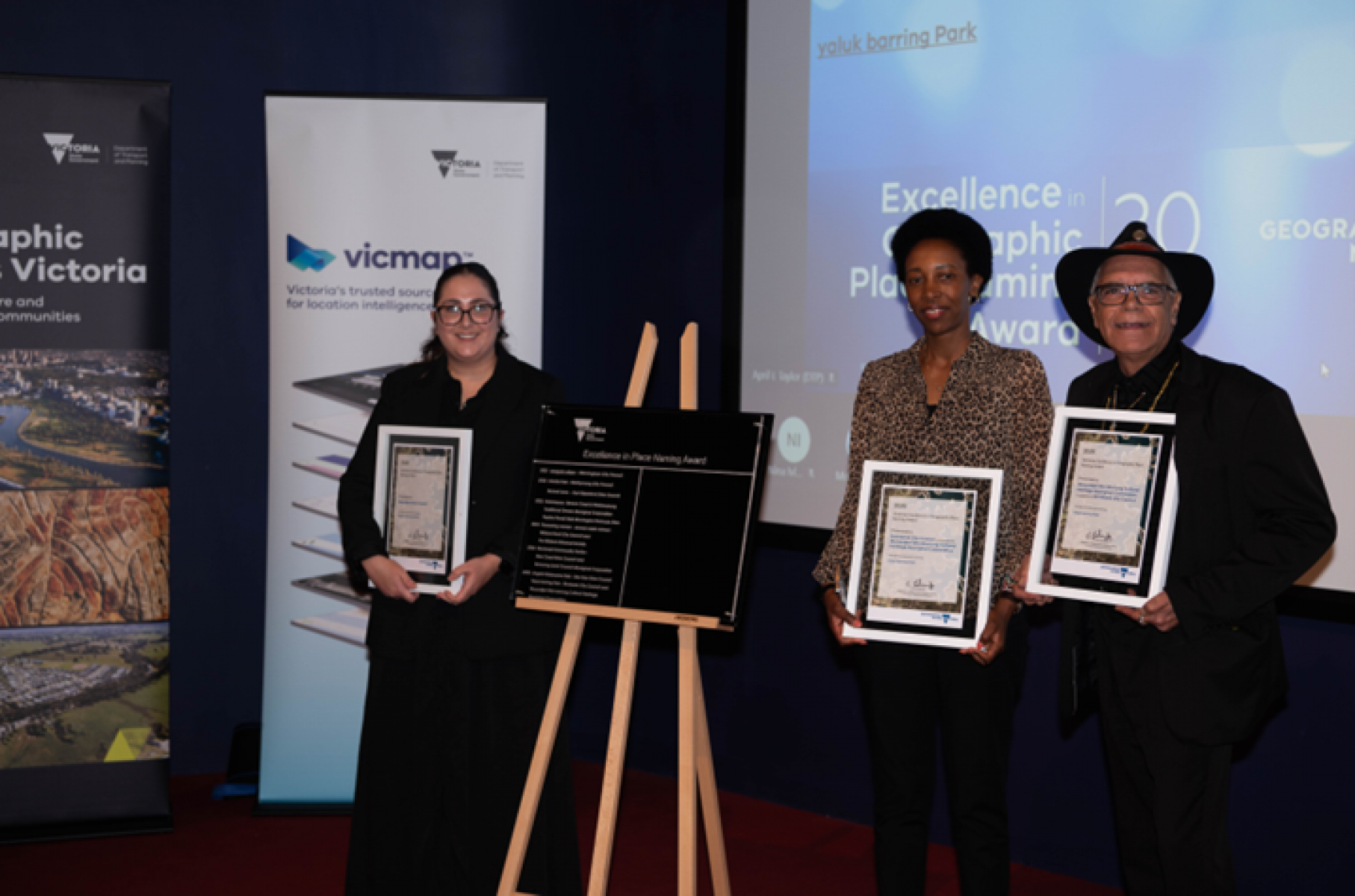 Three people holding framed award certificates, standing around an easel that holds an award plaque. In the background of the photo, banners for Geographic Names Victoria and Vicmap are visible.