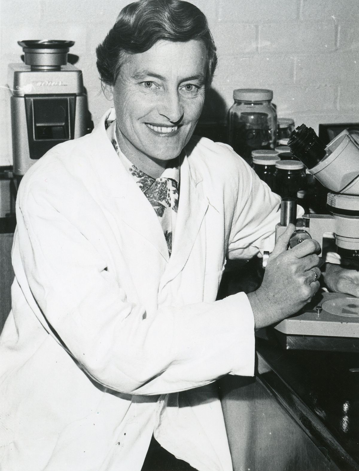 Black-and-white photograph of Dr Isabel ‘Joy’ Bear in a laboratory, seated beside scientific equipment and a microscope while wearing a white lab coat. Dr Bear is holding a laboratory instrument and looking towards the camera. Shelves and jars of research materials are visible in the background.