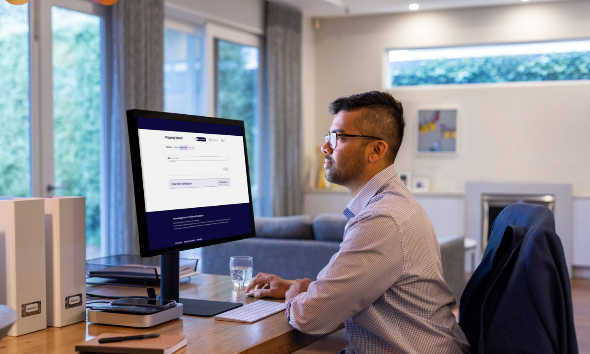 A professionally dressed man sits at a desk looking at a computer monitor with the words 'property search' visible at the top.