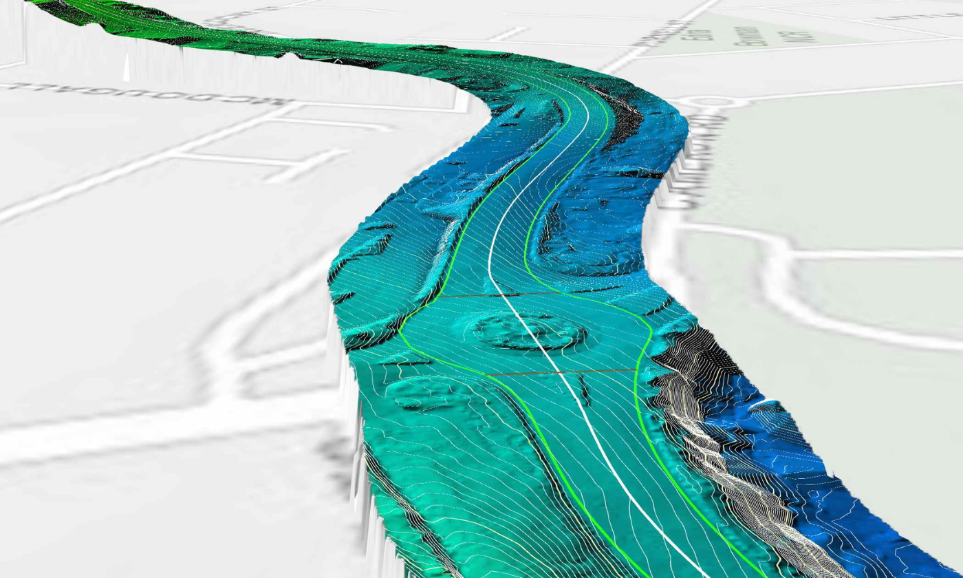 A colourised LiDAR point-cloud image of a section of the great ocean road. The angle tilts diagonally down towards the ground, showing a section of a main road with a roundabout. Details to either side of the road are muted and just light street outlines are visible. Along the main road, and with a buffer of a few metres either side, colour indicates the height of each point, and the details are much more precise. Dark shades of blue represent the deepest points, fading through to shades of green and teal for higher points. A white line indicates the middle of the road, and lime green lines indicate the edges of the road.  
